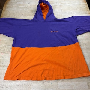 May include: A two-tone hooded sweatshirt in purple and orange. The upper section is purple with an orange interior hood. The lower section is orange. The Champion logo is embroidered in orange on the upper left chest.