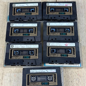 May include: A collection of seven vintage Maxell C90 audio cassette tapes. The black tapes have gold and white labels with handwritten titles like "Elvis Aron Presley" and "KSQY Jazz & Blues #6". The tapes are in clear plastic cases.