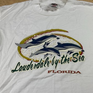 Könnte beinhalten: Ein weißes Kurzarm-T-Shirt mit einer Grafik von zwei Delfinen, die in einem ovalen Rahmen schwimmen. Der Text lautet Lauderdale by the Sea FLORIDA in grüner und roter Schrift. Das Design enthält kleine weiße Blasen und einen Goldrand.