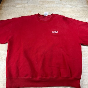 May include: A bright red crewneck sweatshirt featuring long sleeves and a ribbed hem. The front of the garment displays a small, white embroidered Coca-Cola logo with a bottle icon positioned on the upper left chest area.