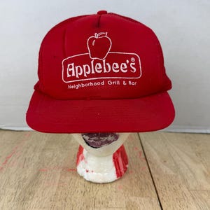 Puede incluir: Gorra de béisbol roja con el logotipo de Applebee's en blanco. La gorra tiene una visera curva y una parte trasera de malla. El texto en la gorra dice "Applebee's Neighborhood Grill & Bar".