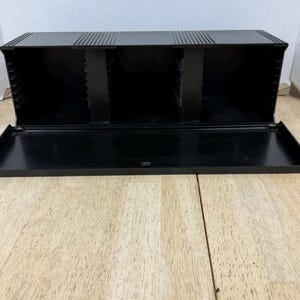 May include: A black plastic storage case with a hinged lid, open to reveal three compartments. The top of the case has a series of horizontal ridges. The case is sitting on a light-colored wooden surface.