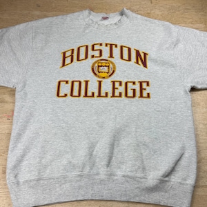 May include: A light grey sweatshirt displaying the words "BOSTON COLLEGE" in maroon lettering. The sweatshirt features a circular emblem in the centre with a gold and maroon design. The garment is made of a soft, comfortable material.