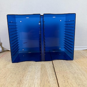 May include: A double-sided blue translucent plastic storage rack featuring multiple horizontal slots on each side for organizing media cases. The unit has a central spine and open-ended compartments designed to hold items vertically in a row.