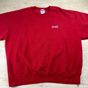 May include: A bright red crewneck sweatshirt featuring a small white Coca-Cola logo embroidered on the upper left chest. The garment has long sleeves, a ribbed collar, and a ribbed hemline. A white brand label is visible inside the neckline.