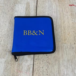 May include: A square blue zippered case featuring the yellow text BB&N printed on the front. The case has a black zipper trim that runs along the edges and a small black zipper pull tab attached to the corner of the closure mechanism.