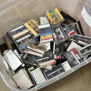 May include: A clear plastic storage bin filled with a collection of assorted audio cassette tapes. The tapes feature various brands, colors, and designs, with some in plastic cases and others with handwritten labels on the paper inserts.