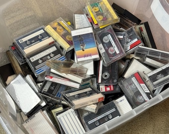 May include: A clear plastic storage bin filled with a collection of assorted audio cassette tapes. The tapes feature various brands, colors, and designs, with some in plastic cases and others with handwritten labels on the paper inserts.