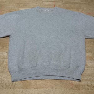 May include: A heather gray crew neck sweatshirt. The sweatshirt has a classic design with a ribbed collar, cuffs, and hem. The fabric appears to be a soft, comfortable material, suitable for casual wear. The garment is laid flat on a wooden surface.