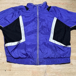 Puede incluir: Chaqueta deportiva vintage morada y negra con cierre de cremallera completo. La chaqueta presenta paneles negros con detalles reflectantes blancos en las mangas y los hombros. El cuello y los puños son acanalados.