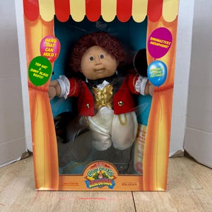 May include: Cabbage Patch Kids Circus Kids Ringmaster doll in its original box. The doll is wearing a red and gold ringmaster outfit with a top hat and shiny black boots. The box has circus-themed graphics and text that reads "Ringmaster's Megaphone!"
