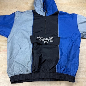 May include: A vintage color-blocked jacket with a hood. The jacket features panels of blue, gray, and black. The black panel has a pocket with the word "MAGIC" in white script. The jacket has a black elastic waistband and a zipper closure.