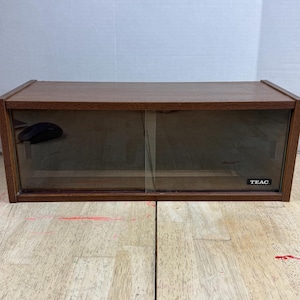 Może przedstawiać: Prostokątna drewniana szafka z przesuwanymi szklanymi drzwiami. Szafka ma ciemnobrązowe wykończenie. Nazwa marki "TEAC" jest widoczna w prawym dolnym rogu. Wewnątrz znajduje się czarna mysz komputerowa.