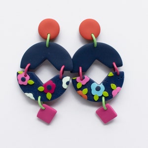 May include: A pair of handmade earrings with a modern design. The earrings feature a navy blue semi-circle shape with a floral pattern, connected to a pink square. The earrings have orange, green, and pink accents.
