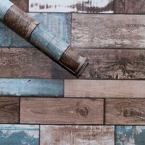 May include: A roll of wood-look self-adhesive wallpaper with a rustic, weathered design. The wallpaper features a mix of brown and blue tones, creating a realistic wood plank effect. The roll is partially unrolled, revealing the adhesive backing.