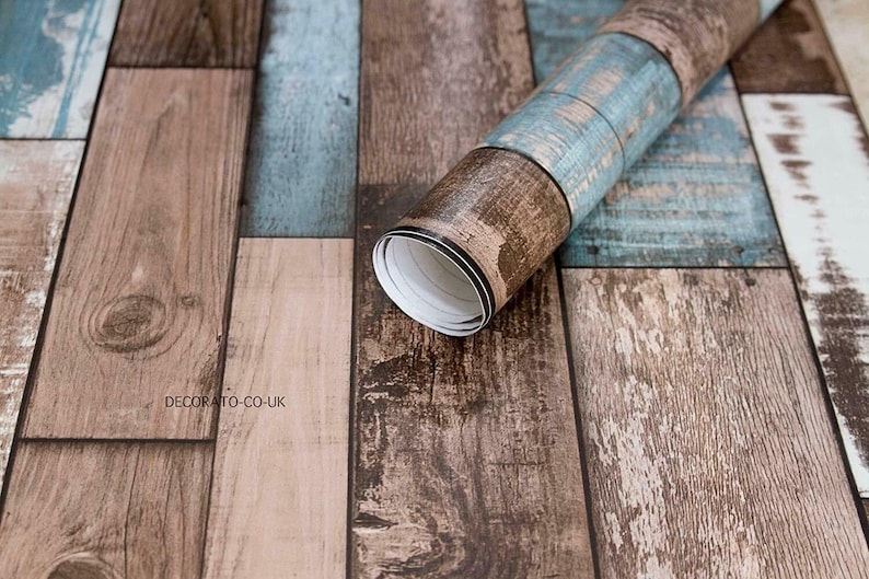 May include: A roll of wood-look self-adhesive wallpaper with a rustic, weathered design. The wallpaper features a mix of brown and blue tones, creating a realistic wood plank effect. The text "DECORATO-CO-UK" is printed on the wallpaper.