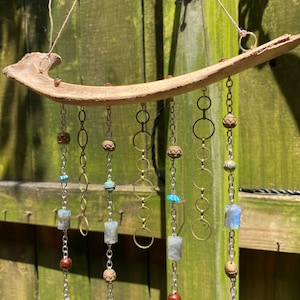 May include: A wooden driftwood wind chime with multiple strands of silver chains and beads. The chains have various charms, including small silver rings, brown wooden beads, and blue and turquoise glass beads. The wind chime is hanging from a wooden surface.