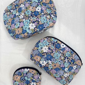 Op de afbeelding: Drie gestikte bloemvormige toilettassen in verschillende maten. De tassen zijn blauw met een wit en roze bloemenpatroon. De grootste tas is bovenaan, de middelste tas is in het midden en de kleinste tas is onderaan.