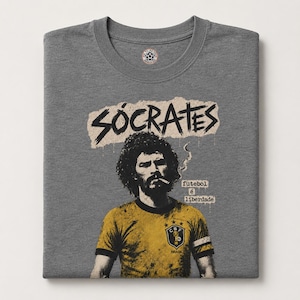 Op de afbeelding: Een grijs gemêleerd t-shirt met ronde hals en een grafische print van voetballer Sócrates. Het ontwerp bevat de naam SÓCRATES in zwarte letters, een portret van de speler met een sigaret en de tekst futebol é liberdade.
