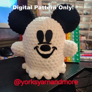 May include: A white crocheted ghost with black felt Mickey Mouse ears, eyes, nose, and a smiling mouth. The ghost is sitting on a dark surface. The text "Digital Pattern Only!" is visible above the ghost. The text "@yorksyarnandmore" is visible below the ghost.