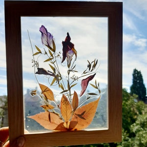 Op de afbeelding: Een houten frame met een heldere glazen inzet met geperste bloemen en bladeren. De bloemen zijn paars, rood en geel, en de bladeren zijn bruin en groen.