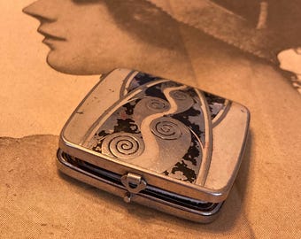 Könnte beinhalten: Ein kleiner, rechteckiger silberfarbener Metall-Kompaktpuder mit einem Jugendstil-Design auf dem Deckel. Das Design zeigt wirbelnde Muster in Blau-, Schwarz- und Silbertönen. Der Kompaktpuder hat einen Klappverschluss.
