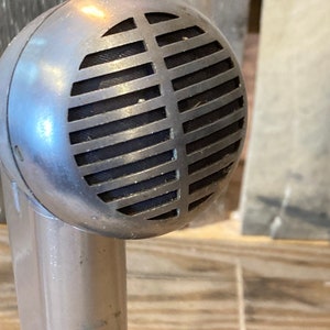 May include: Vintage silver microphone with a round head featuring horizontal grille bars. The microphone has a cylindrical base and is likely used for recording or broadcasting. The design suggests a classic, retro aesthetic.