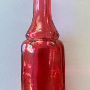 Op de afbeelding: Een vintage, robijnrode glazen fles met een lange hals en een bolvormige basis. De fles heeft een glad, reflecterend oppervlak en is leeg. De fles wordt waarschijnlijk gebruikt voor het bewaren van vloeistoffen, zoals parfum of medicijnen.