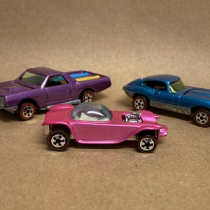 May include: Three vintage toy cars in purple, pink, and blue. The purple car is a pickup truck with a yellow stripe. The pink car has a bubble top. The blue car is a sports car. All have red-lined tires.