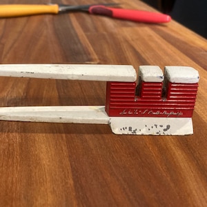 May include: A vintage knife sharpener with a white handle and red sharpening component. The sharpener has the text "Lib-Sharp" on the red section. Two knives are in the background, one with a yellow handle and the other with a red handle.
