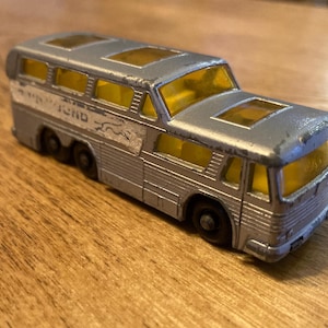 May include: A vintage silver toy bus with yellow tinted windows and roof panels. The bus has six wheels and the word "FORD" is visible on the side. The toy bus is sitting on a wooden surface.