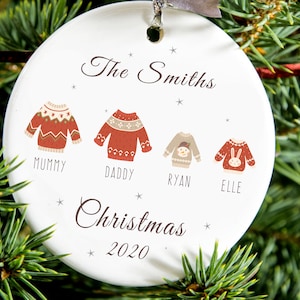 May include: A white ceramic Christmas ornament with a family name and the text "Christmas 2020." The ornament features four sweater designs, each labeled with a family member's name. The ornament is hanging from a silver ribbon.