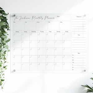 May include: A clear acrylic monthly planner with the text "The Jacksons' Monthly Planner." The planner features a calendar grid, a shopping list section, and a notes section. It is mounted on a white wall with silver hardware.
