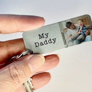 May include: A rectangular metal keychain with the words "My Daddy" printed on the left side. The right side features a color photograph of a man and a child. The keychain is attached to a keyring with several keys.