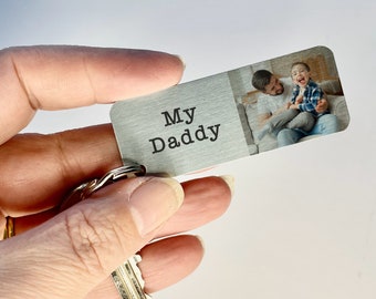 May include: A rectangular metal keychain with the words "My Daddy" printed on the left side. The right side features a color photograph of a man and a child. The keychain is attached to a keyring with several keys.