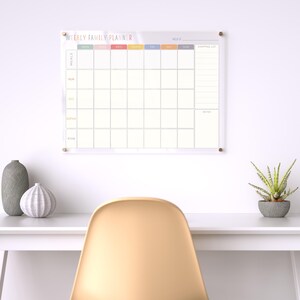 Pu&ograve; includere: Un pianificatore settimanale familiare in acrilico trasparente con la scritta "WEEKLY FAMILY PLANNER" in lettere colorate. Il pianificatore ha sezioni per i pasti, i membri della famiglia e una lista della spesa. &Egrave; montato su un muro bianco.