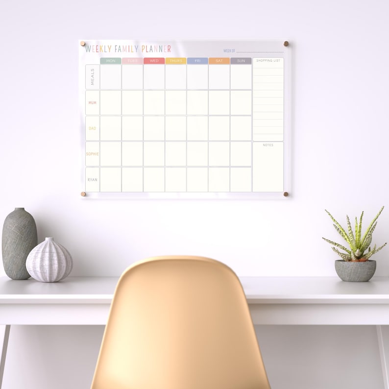 Pu&ograve; includere: Un pianificatore settimanale familiare in acrilico trasparente con la scritta "WEEKLY FAMILY PLANNER" in lettere colorate. Il pianificatore ha sezioni per i pasti, i membri della famiglia e una lista della spesa. &Egrave; montato su un muro bianco.