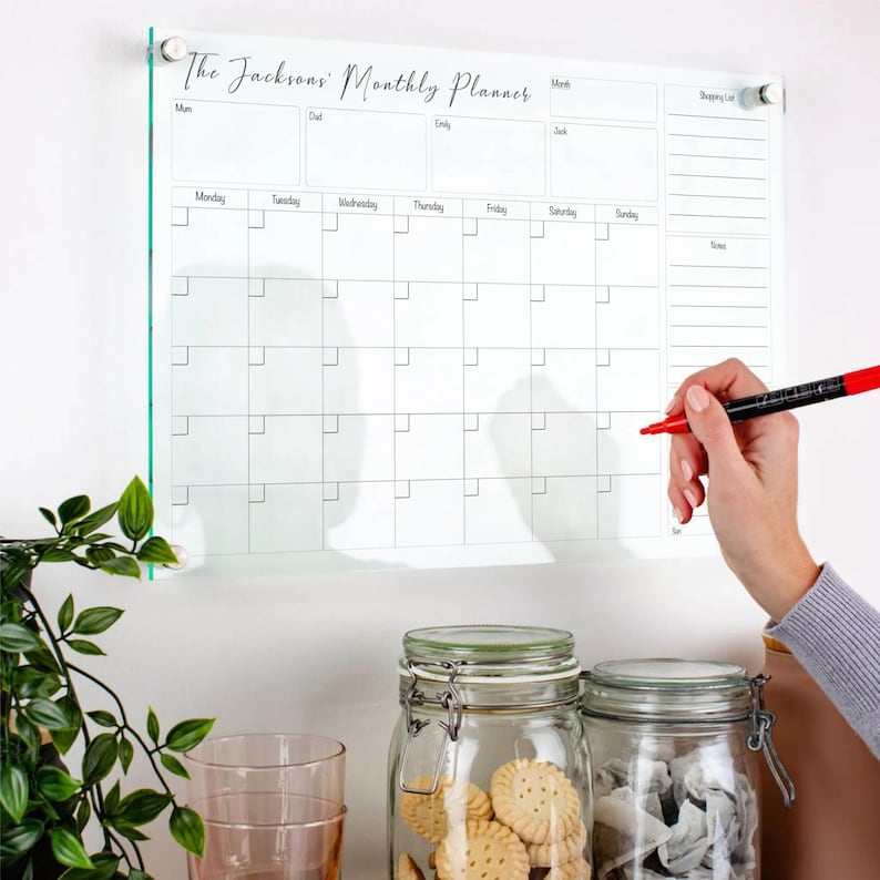 May include: A clear acrylic monthly planner with the text "The Jacksons Monthly Planner" in cursive. The planner has a calendar grid, a shopping list section, and a notes section. A person is holding a red marker near the calendar.