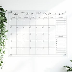 May include: A clear acrylic monthly planner with the text "The Beverland Monthly Planner." The planner has a grid layout for each day of the week, a shopping list section, and a weekly menu section. It is mounted on a white wall.