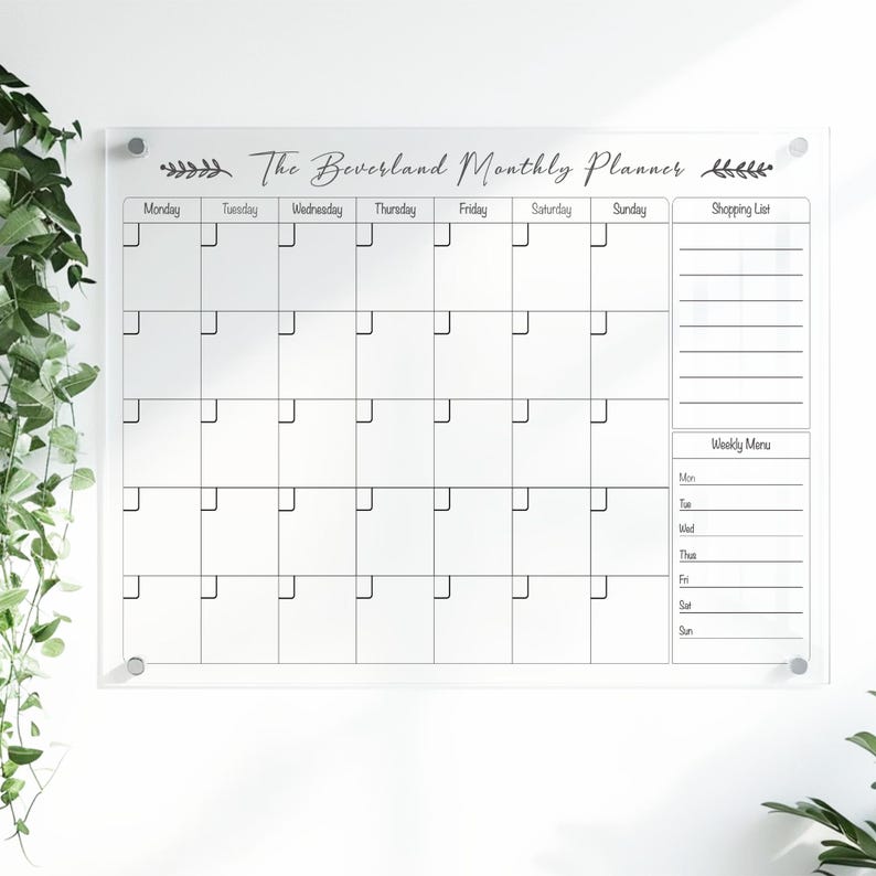 May include: A clear acrylic monthly planner with the text "The Beverland Monthly Planner." The planner has a grid layout for each day of the week, a shopping list section, and a weekly menu section. It is mounted on a white wall.