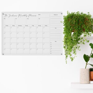 May include: A clear acrylic monthly planner with the text "The Jacksons Monthly Planner" in cursive script. The planner includes sections for the month, shopping list, notes, and a calendar grid. It is mounted on a white wall with a hanging plant.