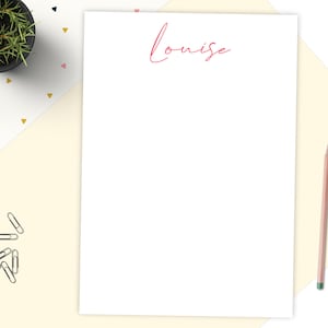 May include: A white sheet of stationery with the name "Louise" in red script. A pencil, a small potted plant, and paper clips are arranged on a cream and white surface with geometric shapes.