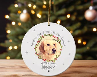 May include: A white ceramic Christmas ornament with a photo of a golden retriever dog. The ornament has text that reads "Leaving Paw Prints On Our Hearts at Christmas Benny" and is decorated with a wreath and star accents. A gold string is attached for hanging.