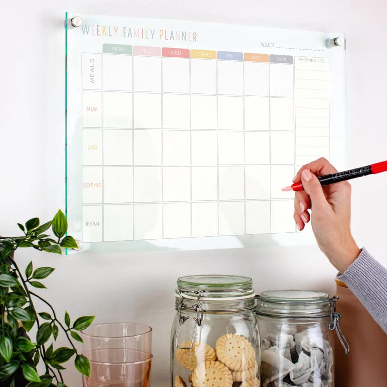 Pu&ograve; includere: Un'agenda settimanale familiare in vetro trasparente con un'intestazione colorata. L'agenda &egrave; montata su una parete bianca e include sezioni per i pasti e una lista della spesa. Una mano tiene in mano un pennarello rosso, pronta a scrivere sull'agenda.