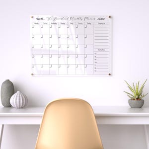 May include: A clear acrylic monthly planner with the text "The Beversland Monthly Planner" and a grid layout for scheduling. The planner is mounted on a white wall with gold-colored hardware. A white desk with decorative items and a chair are in the foreground.