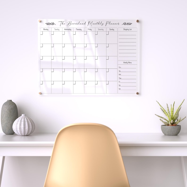 May include: A clear acrylic monthly planner with the text "The Beversland Monthly Planner" and a grid layout for scheduling. The planner is mounted on a white wall with gold-colored hardware. A white desk with decorative items and a chair are in the foreground.