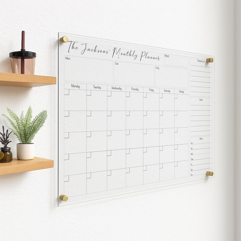 May include: A clear acrylic monthly planner with gold-colored mounting hardware. The planner features a grid layout for daily scheduling, a shopping list section, and the text "The Jacksons' Monthly Planner" in elegant script.
