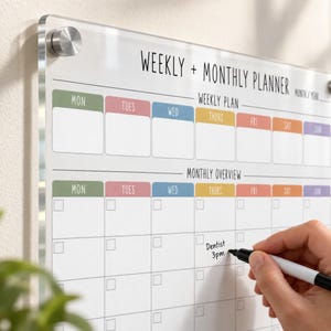 May include: A clear acrylic wall-mounted weekly and monthly planner board with silver metal standoffs. The board features printed black text, colourful day headers, and grid sections for scheduling. The text reads Weekly + Monthly Planner.
