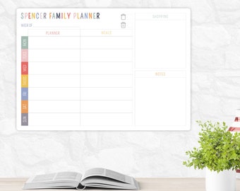 May include: A white family planner with the text "SPENCER FAMILY PLANNER" in colorful letters. The planner includes sections for each day of the week, planner, meals, shopping, and notes. A small green plant and an open book are in the foreground.