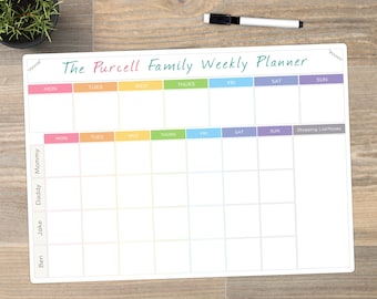 May include: A white weekly family planner with the text "The Purcell Family Weekly Planner" in green script. The planner has color-coded days of the week and spaces for each family member. A black and white marker and a small potted plant are also visible.